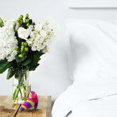 A wooden bedside table holds a vase filled with white flowers and green foliage. Next to the vase, there is the Avant Free Spirit Vibrating Anal Plug in Scarlet by Avant, featuring a colorful, abstract pattern and 10 vibrating functions, discreetly tucked in. A white bed with neatly arranged pillows sits adjacent to the table, set against a blank white wall in the background.